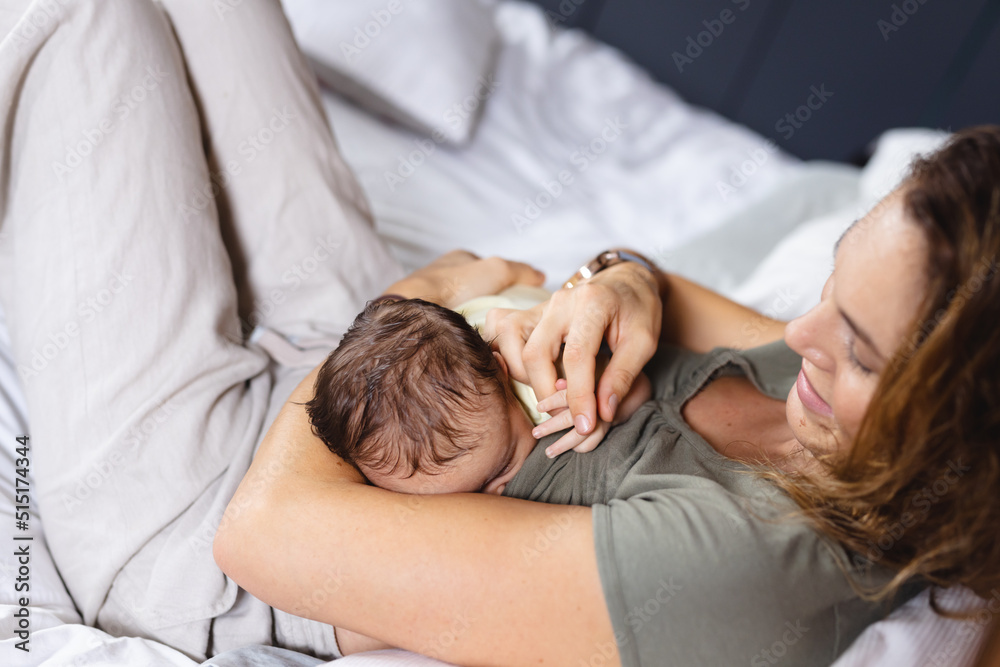 Happy caucasian mid adult mother breastfeeding newborn baby on bed at home, copy space