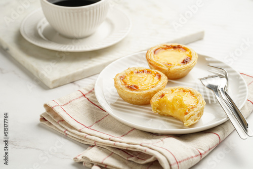 Traditional portuguese vanilla pudding puff pastry pastel de nata on black oven rack on white plate on checkered kitchen towel with a cup of coffee in white porcelain