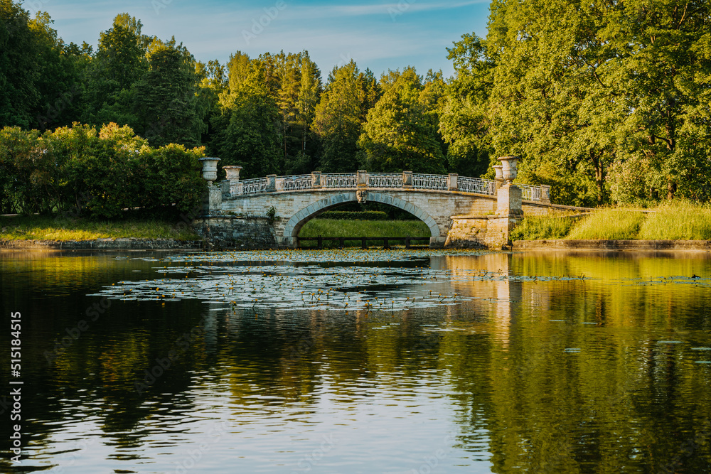 View of the vintage  Viscontiev Bridge and Slavyanka River in the Pavlovsk Palace and Park Complex on a summer sunny day. Water lilies bloom on the river.
