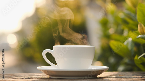 Natural steam smoke of coffee from a white hot coffee cup on an old wooden table with wonderful morning warm sunshine flare nature view outside. Coffee cup, hot drink, espresso, and breakfast concept