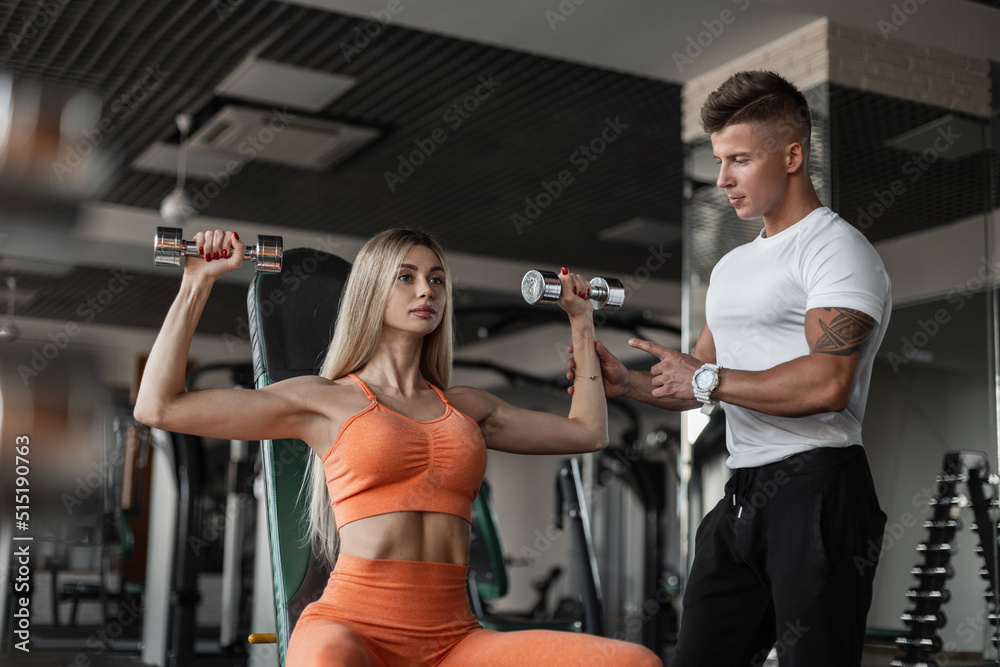 Handsome bodybuilder trainer man with a tattoo trains a beautiful blonde woman and points out how to do exercises in the gym. Sporty girl workout with dumbbells