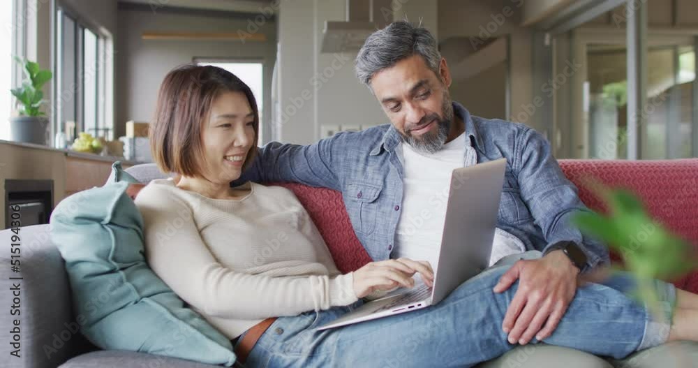 Happy diverse couple sitting in living room using laptop and talking Happy diverse couple sitting in living room using laptop and talking