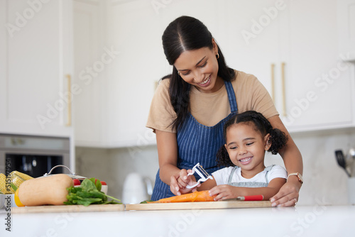 Caring mother and adorable daughter cooking and bonding at home. Happy little girl helping her mother while using peeler and preparing vegetables for a healthy meal together photo
