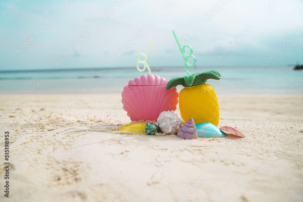 Exotic tropical summer drinks in pink shell shaped and pineapple shaped bottle with colorful sea shells on the sandy beach background, Maldives