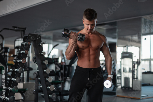 Sporty strong handsome male bodybuilder with a naked muscular athletic torso training and workout with dumbbells in the gym