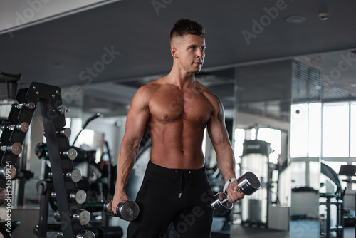 Cool handsome young male bodybuilder athlete with a sporty muscular naked torso working out in the gym with dumbbells