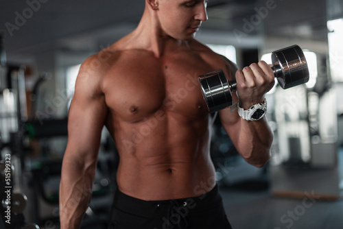 Handsome male athlete with a muscular fitness body holding a metal dumbbell and pumping his biceps muscles in the gym