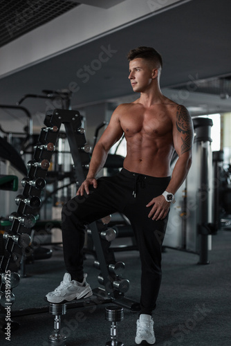 Handsome young athletic guy with hair and a bare muscular torso stands and poses near the dumbbells in the gym