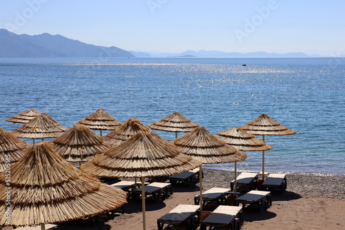 Fototapeta Naklejka Na Ścianę i Meble -  Empty pebble beach with wicker parasols and deck chairs. Picturesque view to blue sea and mountains in mist, luxury summer resort