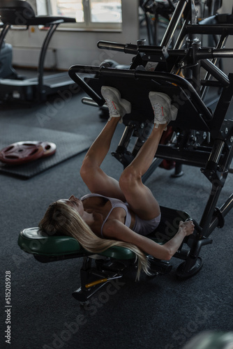 Beautiful athletic strong girl with a sexy body in trendy sports clothes with sneakers is lying on the simulator and working out her leg muscles in the gym