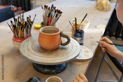 Boy coloring clay cup on a potter's wheel. Teaching pottery is Healthy free time for children. Kid painting ceramic cup on a wooden table at workshop. Hobbies, craft and handwork