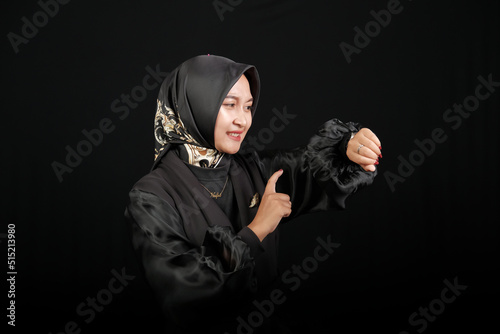 Happy Asian Muslim woman in a black dress wearing glasses looking at her watch and smiling against a black screen background