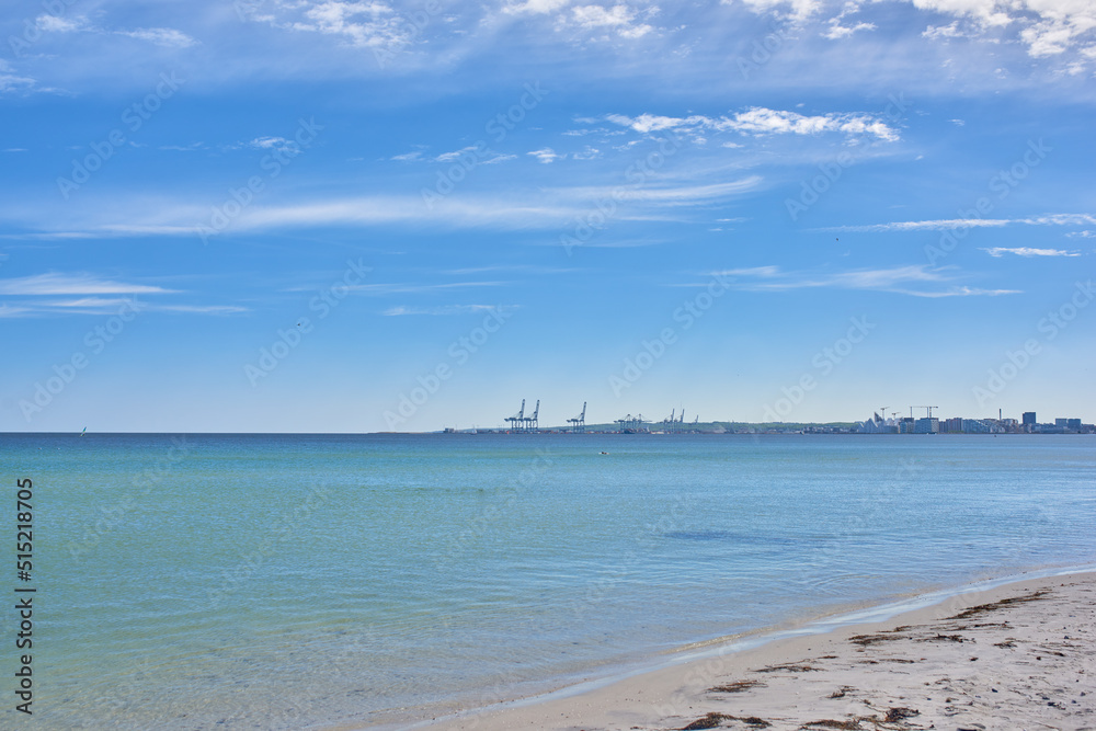 Obraz premium Copy space at the sea with a blue sky background. Calm ocean waves wash onto stones at an empty beach shore with a port or harbor on the horizon. a scenic landscape for a relaxing summer holiday