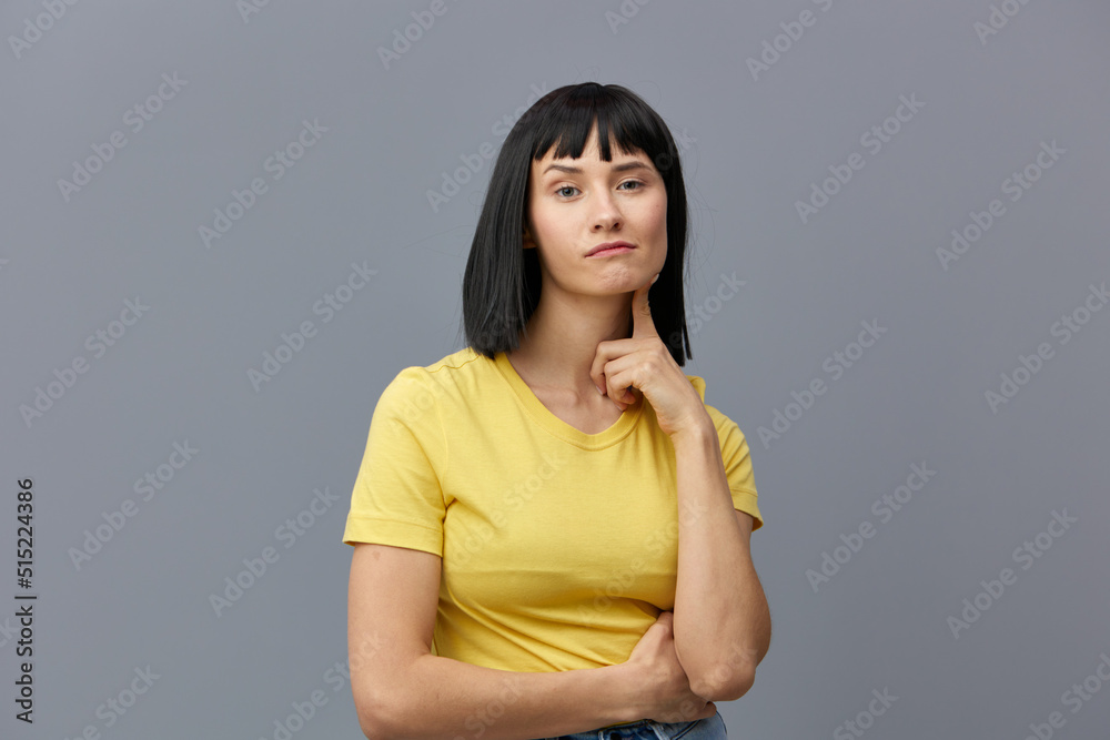 a beautiful, attractive playful woman poses standing on a gray background in a yellow T-shirt and playfully looking at the camera with her index finger touching her face a beautiful, attractive playful woman poses standing on a gray background in a yellow T-shirt and playfully looking at the camera with her index finger touching her face