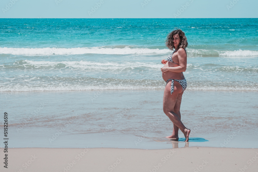 Happy pregnant woman on the beach. High risk pregnant adult lady smiles on the beach. Miss in a bikini with a big fat belly of eight months. Overweight and health concept enjoying her unborn baby Happy pregnant woman on the beach. High risk pregnant adult lady smiles on the beach. Miss in a bikini with a big fat belly of eight months. Overweight and health concept enjoying her unborn baby