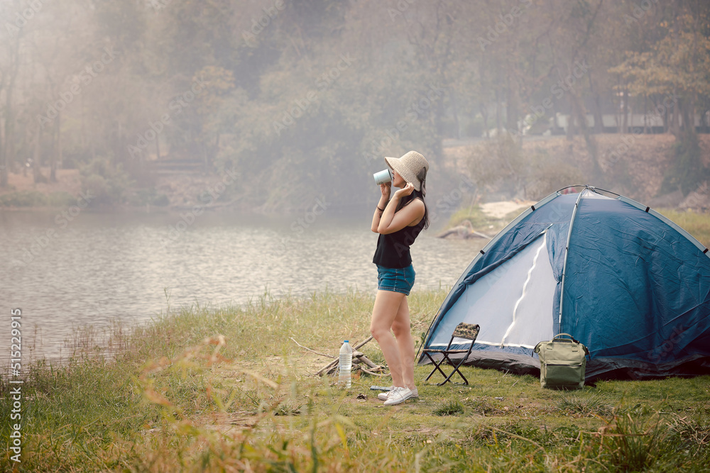 Happy woman drinking coffee or tea in front of the camping tent at meadow near lake. Recreation and journey outdoor activity lifestyle. Travel and adventure theme. Female tourist portrait.