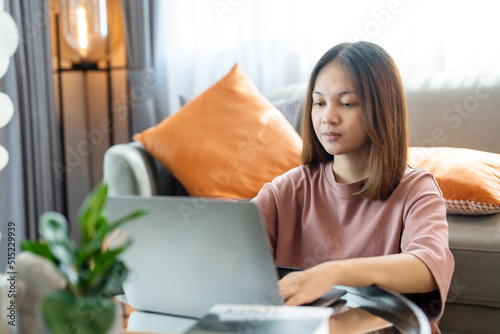 young Asian woman is studying online in front of her laptop computer and typing a message. She showed signs of thinking by using her left hand to support her hold. Work from home.