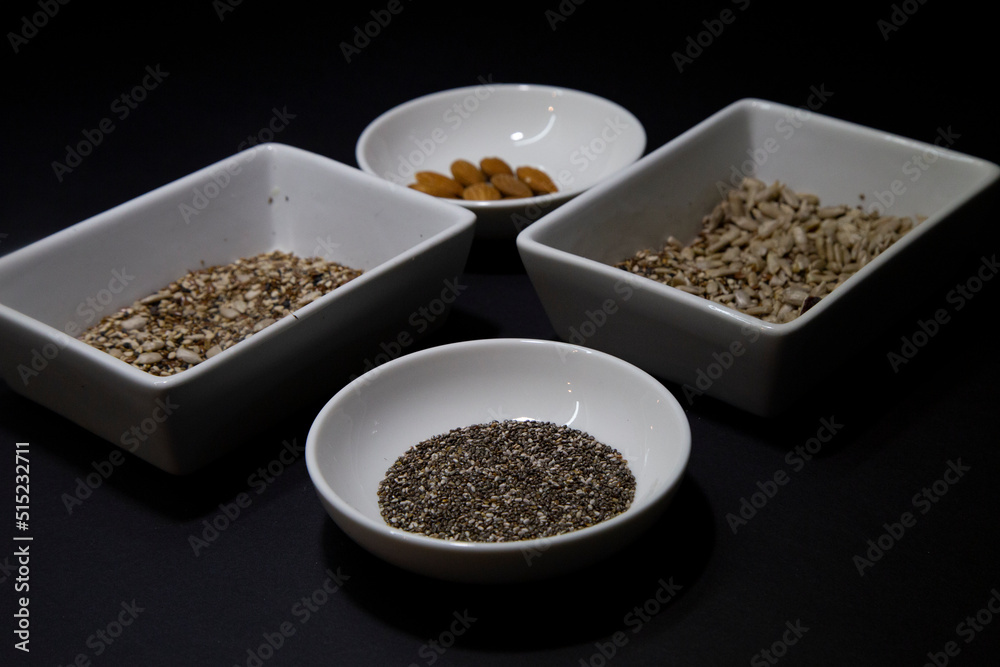 Almonds, chia and other healthy vegan seeds in white porcelain bowls on a black background Almonds, chia and other healthy vegan seeds in white porcelain bowls on a black background