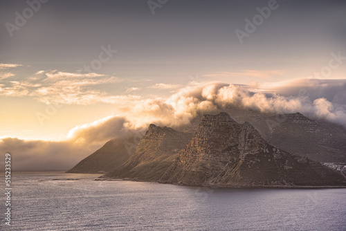 Panorama of a mountain coastline with a cloudy sunset in South Africa. Scenic landscape of soft white clouds covering Table Mountain at dusk near a calm peaceful sea at Hout Bay near Cape Town.