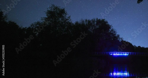 The movement of stars in the night sky over the bridge with colored backlighting, timelapse of the night landscape. Silhouette of trees. photo