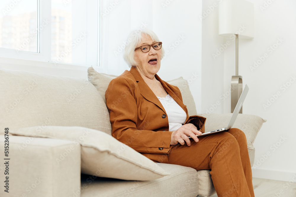 a happy, pleasantly shocked elderly woman is sitting on a sofa in a bright interior and looks at the camera with her mouth wide open, holding a laptop on her lap a happy, pleasantly shocked elderly woman is sitting on a sofa in a bright interior and looks at the camera with her mouth wide open, holding a laptop on her lap