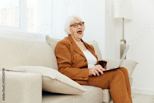 a happy, pleasantly shocked elderly woman is sitting on a sofa in a bright interior and looks at the camera with her mouth wide open, holding a laptop on her lap © Tatiana