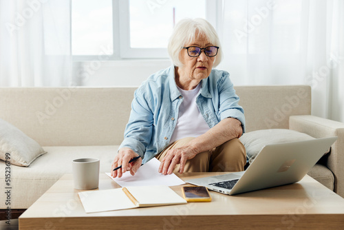 a pleasant elderly lady, passionate about work, sits in her apartment on a comfortable sofa in stylish clothes and uses a laptop and looks at her notebook for notes