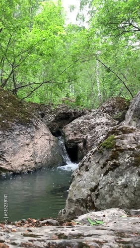 Mountain river - a small waterfall on a river with crystal clear water that flows among gray stones in a green forest on a cloudy summer day. Big stones near the pond