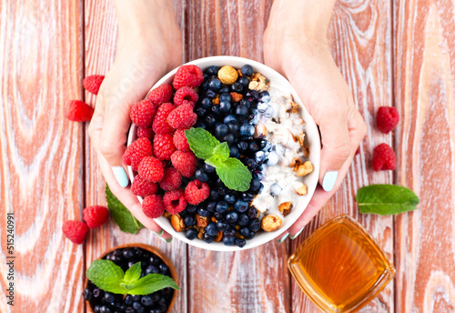 The girl's hands hold a bowl of muesli with ripe berries, yogurt and honey on the wooden table. Healthy breakfast. Top view. Сlose-up. Selective focus.