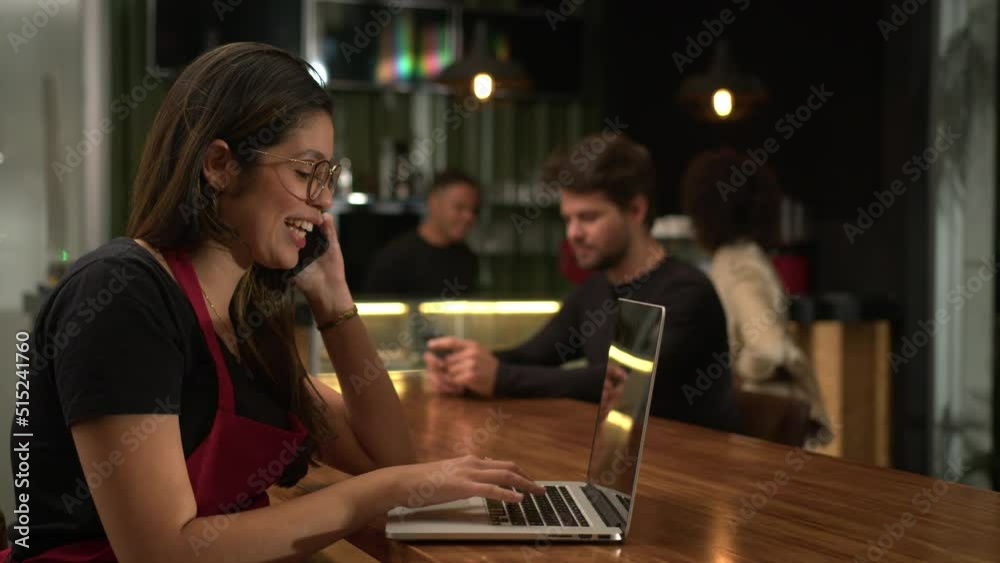 Young woman staff talking on phone and receiving online orders in front of laptop computer sitting at coffee shop Young woman staff talking on phone and receiving online orders in front of laptop computer sitting at coffee shop