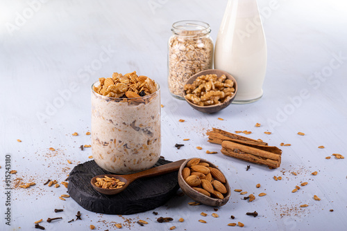 Overnight oats with Granola. Accompanied with almonds and a spoon of oats. Cloves and cinnamon scattered. Cinnamon whole. In the background nuts, milk, and raw oats.