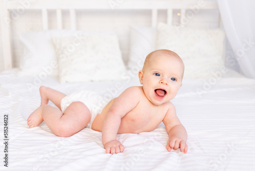 laughing baby girl in diapers in a crib on a white cotton bed lying on her stomach in the nursery smiling, newborn morning