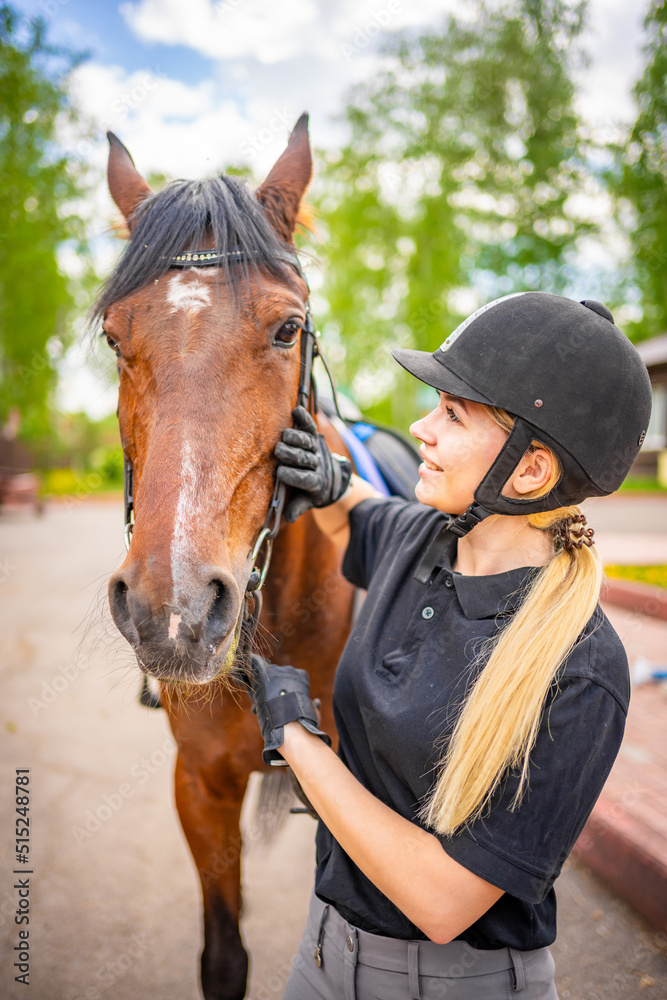 Fototapeta premium Lovely young woman wearing helmet stroking to her brown horse