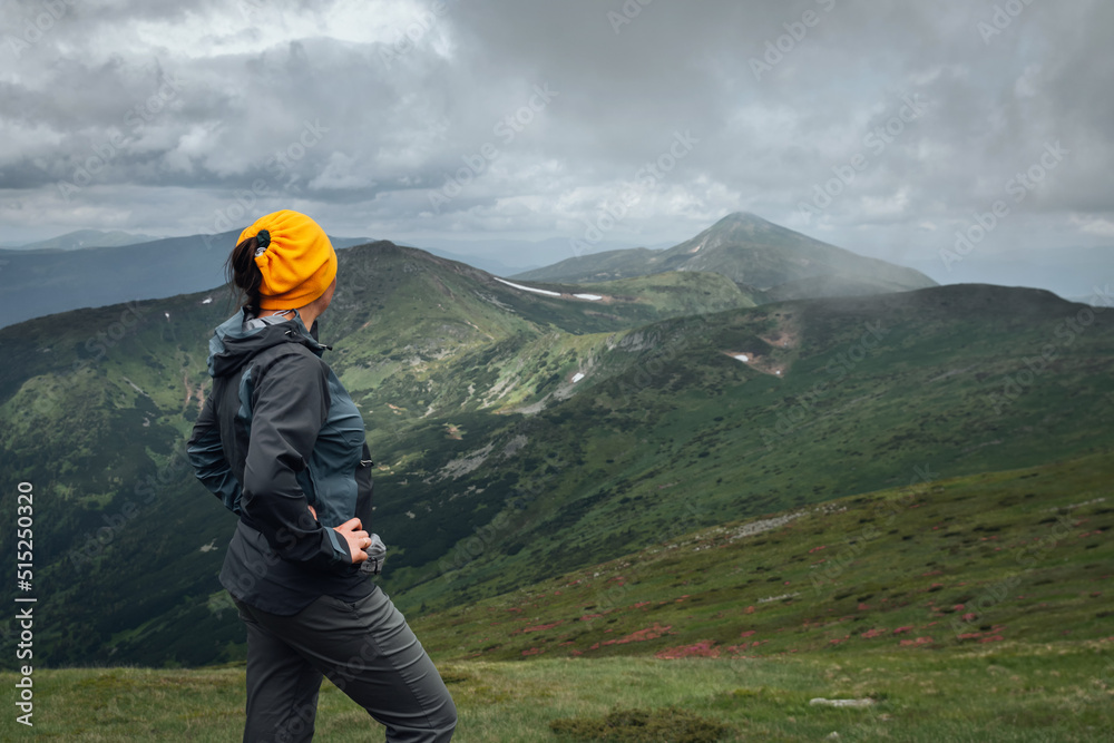 Girl hiker stands on a rock in the mountains. Trekking life. Hike through the Carpathian mountains. Green mountain slopes and blooming rhododendron. Girl hiker stands on a rock in the mountains. Trekking life. Hike through the Carpathian mountains. Green mountain slopes and blooming rhododendron.