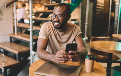 Happy African American male blogger in classic eyewear looking away and smiling while creating content plan in coworking space, cheerful hipster guy with modern cellular technology enjoying freelance