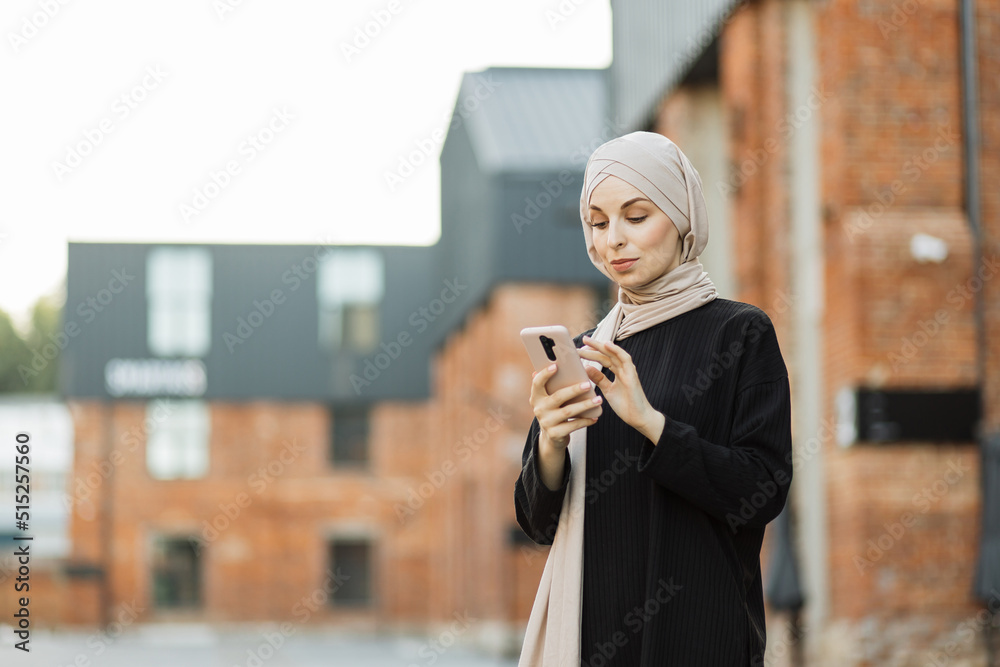 Fototapeta premium Attractive smiling muslim woman in hijab texting on smart phone standing on background of buildings in city street. Portrait of young arabian businesswoman using mobile phone in city.