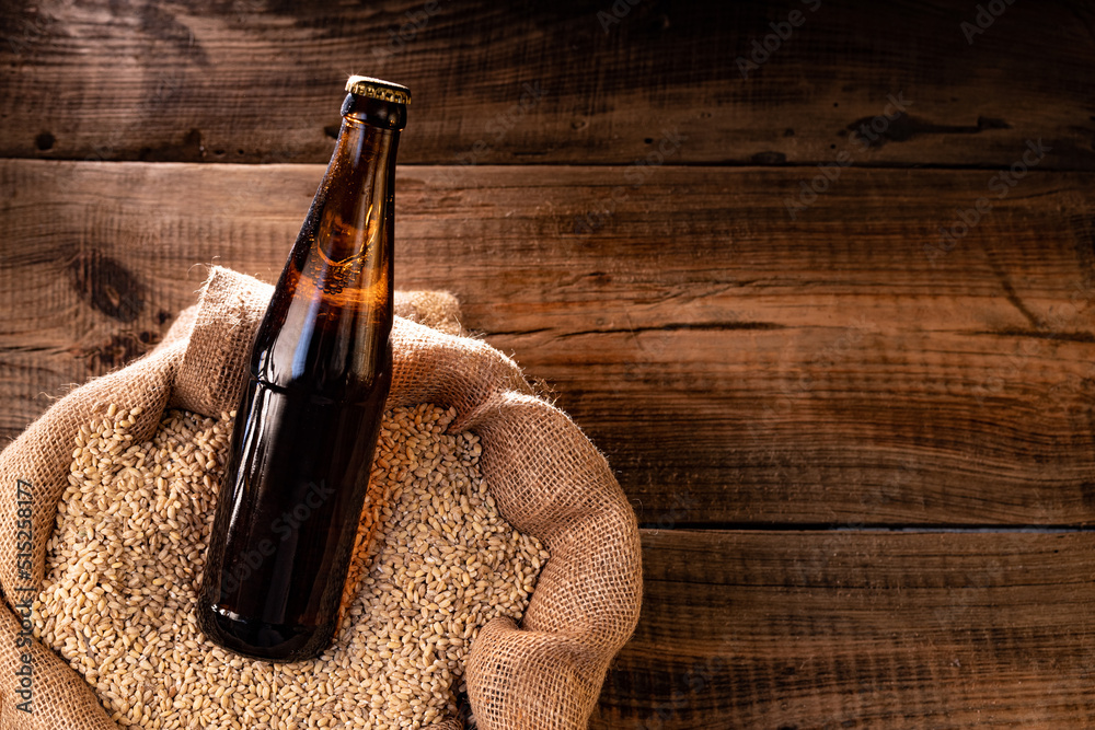 A bottle of dark beer in a bag of wheat. A beam of light falls on a bottle of beer. View from above. - obrazy, fototapety, plakaty 