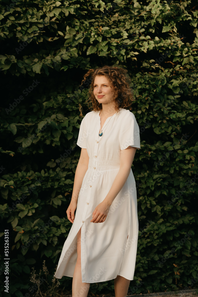 A happy woman with curly hair is posing in the garden in the evening. A lady in a white dress with green leaves in the background. A happy woman with curly hair is posing in the garden in the evening. A lady in a white dress with green leaves in the background.