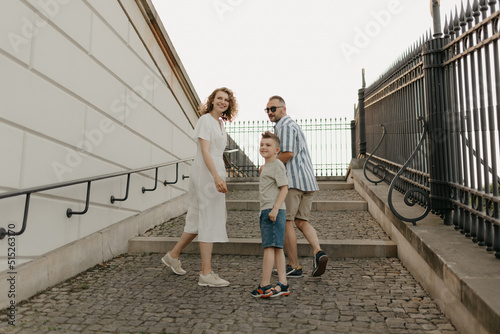 A photo from the back of a family is looking back while climbing the stairs of the palace in an old European town. A happy father, mother, and son are holding hands in the evening.