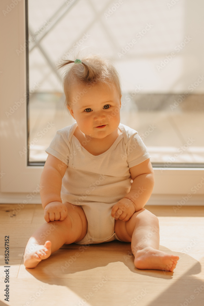 A kind 7-month girl is sitting near a balcony door in a bodysuit at home. A cute infant is waiting for her parents in the sunlight. A kind 7-month girl is sitting near a balcony door in a bodysuit at home. A cute infant is waiting for her parents in the sunlight.