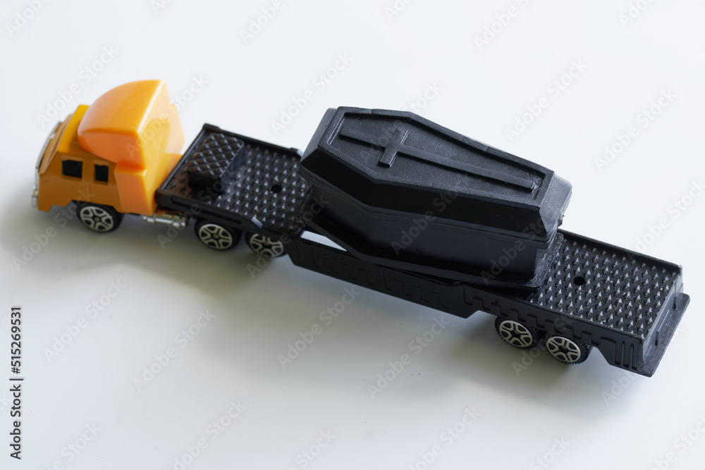 Yellow toy truck with a trailer transports a black coffin. White background. The concept of transporting the dead and transporting corpses. Toy world. Selective focus.