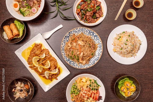 Set of asian dishes seen from above, lemon breaded chicken fillets, three rice delicacies, stir fry noodles, spring rolls, takoyaki, pork ramen and poke bowl, soy sauce on dark wooden table