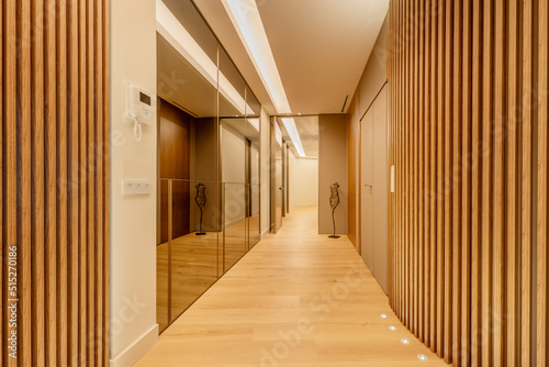 Hallway of a house with a wall covered with mirrors on one side and wooden slats on the other, chestnut floorboards and recessed lights in the floor