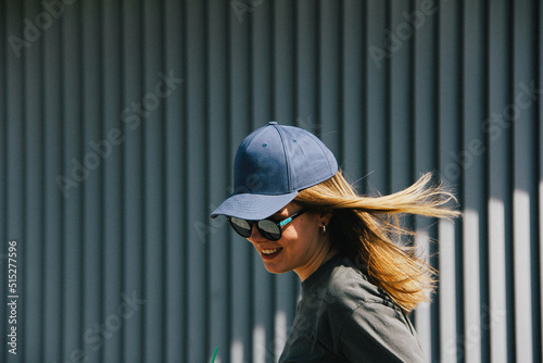 a girl in a cap and sunglasses smiles against a gray ribbed wall