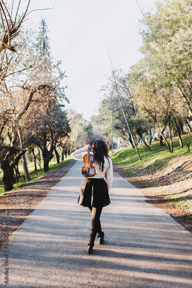curly brunette woman holding a violin on her back, walking through the park road at sunset. curly brunette woman holding a violin on her back, walking through the park road at sunset.