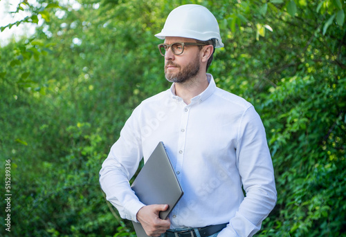 Builder man in hardhat holding laptop outdoors