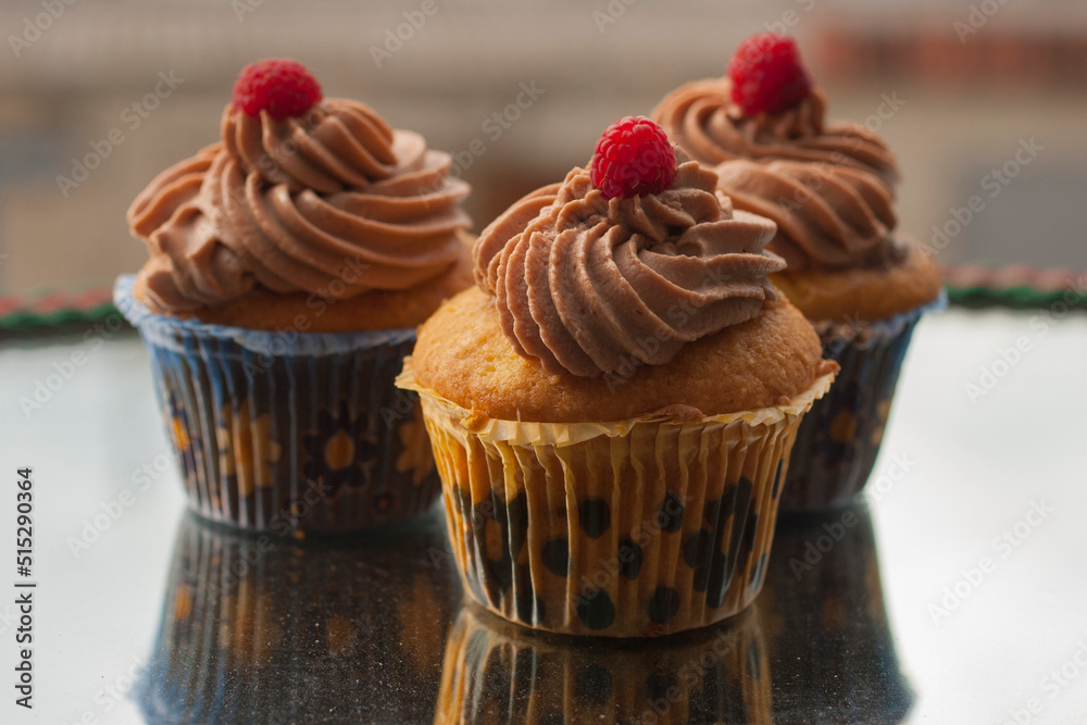 Beige cupcakes made of delicate light sponge cake decorated with chocolate cream. Topped with ripe raspberries.