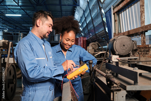 Asian male and female African American engineers in safety uniform work by inspecting machines' voltage current, checking, and maintaining at manufacture factory, electric system service occupations.