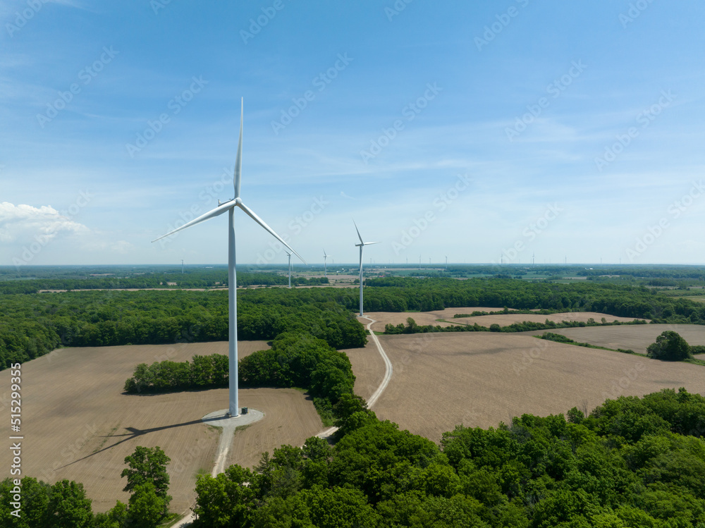 A wind turbine is seen in a rural landscape, on a partly cloudy and sunny day. Multiple wind turbines can be seen in the far landscape.