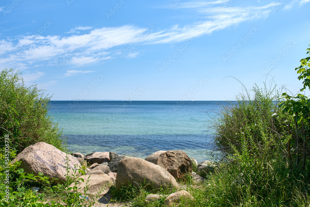 A shallow rocky coast on a calm quiet beach day during summer with grass growing on the shore. Scenic view of a crystal blue ocean with clear blue skies and white clouds during a warm summer day